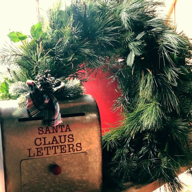 A vintage metal mailbox stamped 'Santa Claus Letters' tucked into a fresh evergreen wreath with a plaid bow
